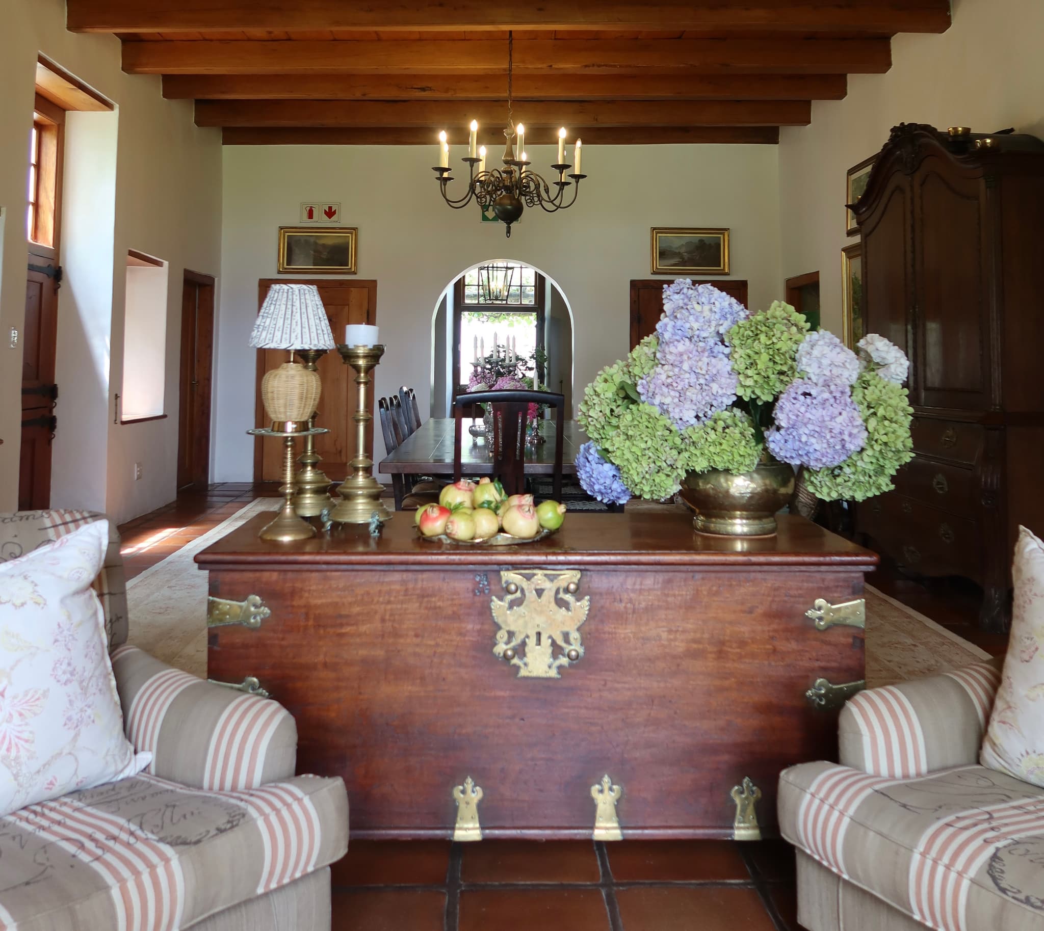 Entrance hall with antique wooden chest, hydrangeas, brass candlesticks, and striped armchairs